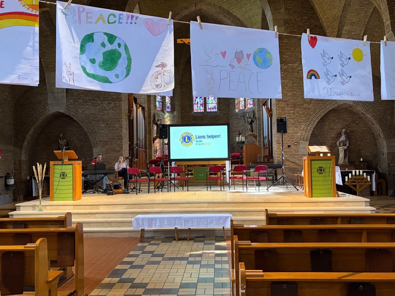 Kindertekeningen met "Peace", duiven, harten en de wereldbol hangen aan een lijn boven het podium in een kerk; op een scherm staat "Lions helpen! Raalte".