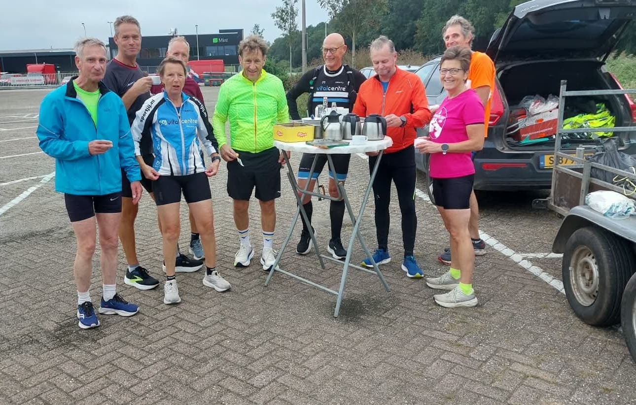 Hardlopers en fietsers staan in sportkleding rond een statafel met thermoskannen en koffie op een parkeerplaats.