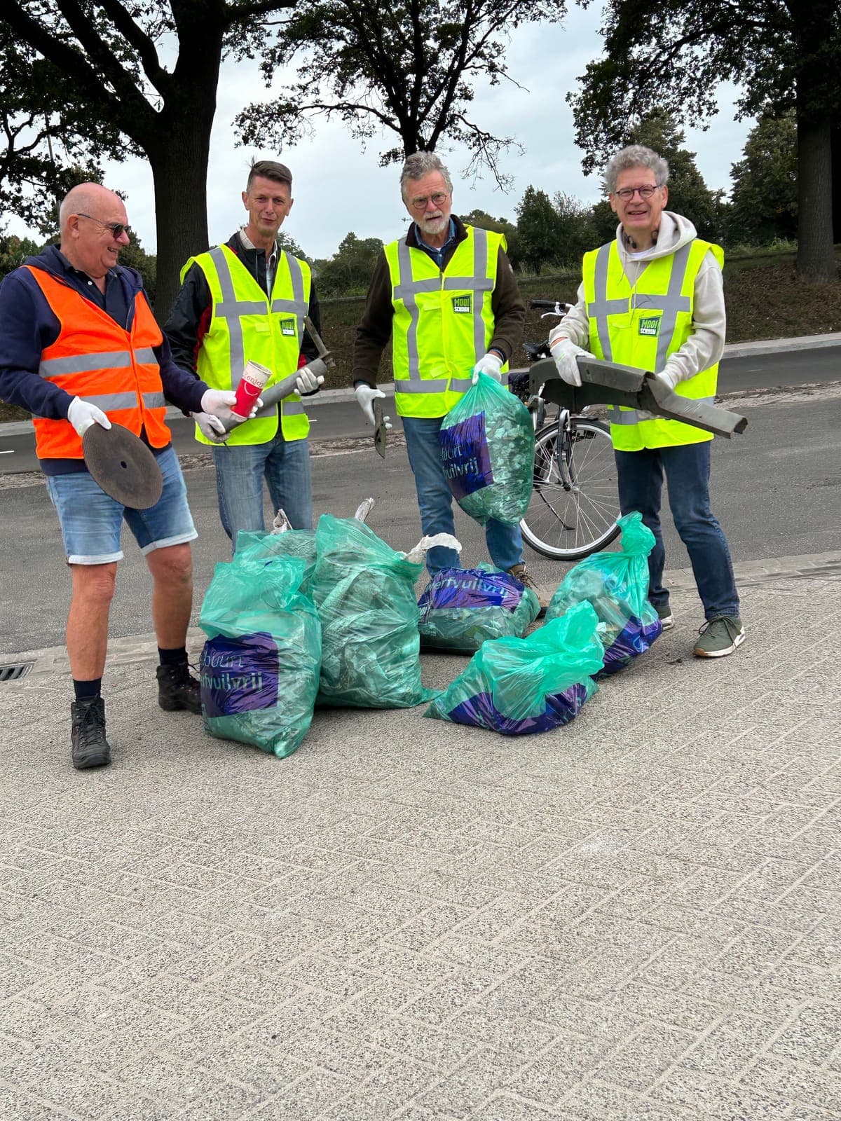 Vier vrijwilligers staan bij een stapel volle vuilniszakken langs de weg en houden gevonden fietsonderdelen en stukken metaal omhoog.