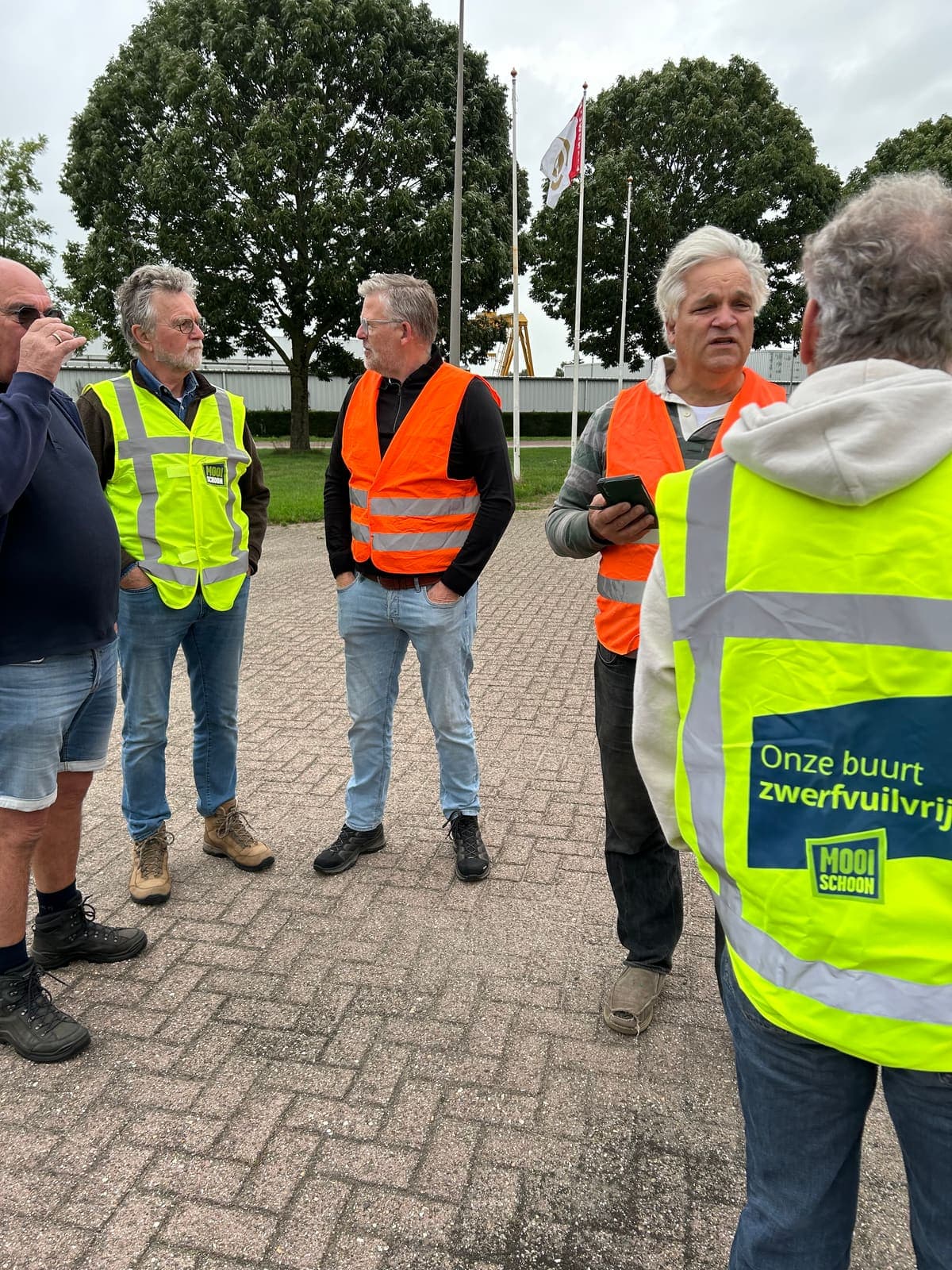 Een groep deelnemers in veiligheidshesjes staat op een plein te overleggen voor de start; op een hesje staat "Onze buurt zwerfvuilvrij".
