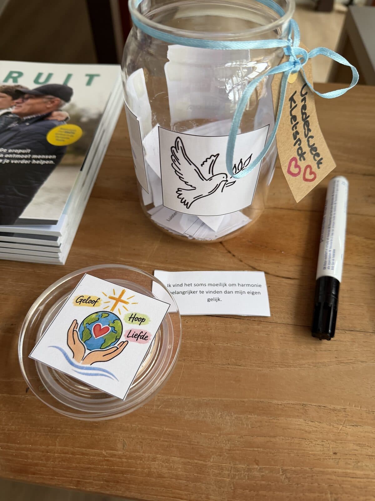 A glass jar with a dove on it and the label "Vredesweek kletspot", full of notes, with a marker and a card reading "Geloof, Hoop, Liefde" beside it.